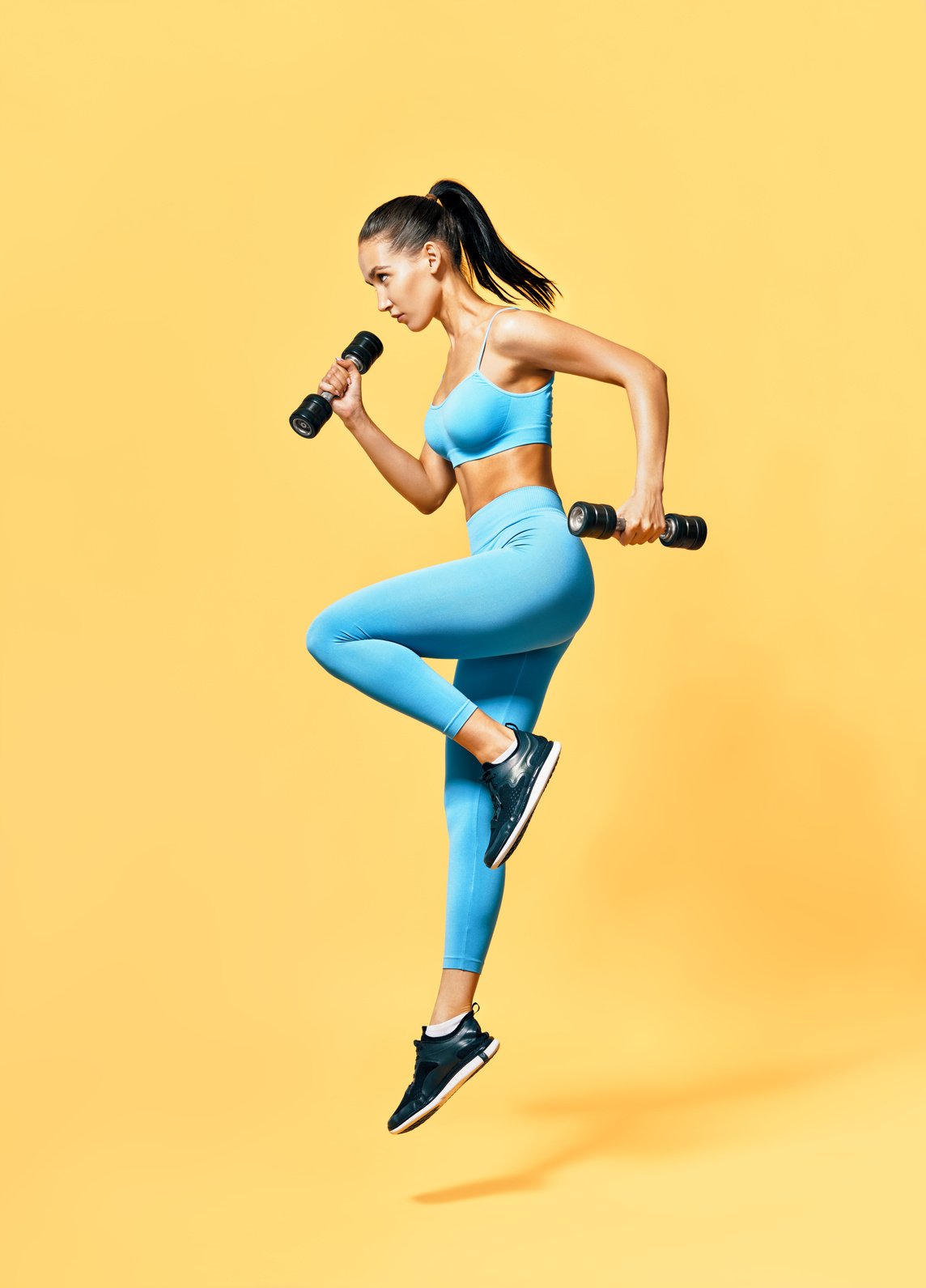 Woman in Sportswear Jumping with Dumbbells on Yellow Background 